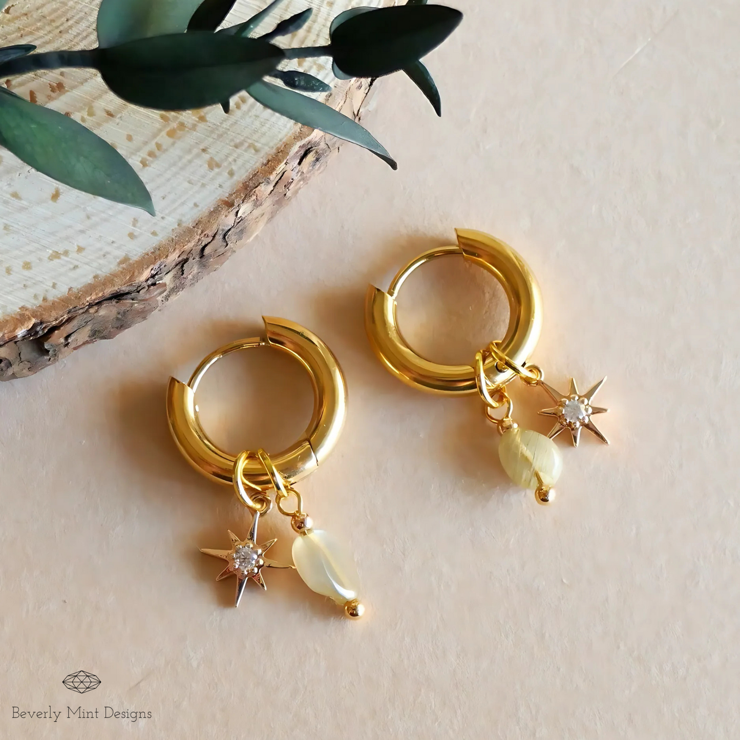 A pair of gold hoop earrings with star-shaped charms and a small gemstone dangling from them, placed on a wooden surface alongside some greenery.