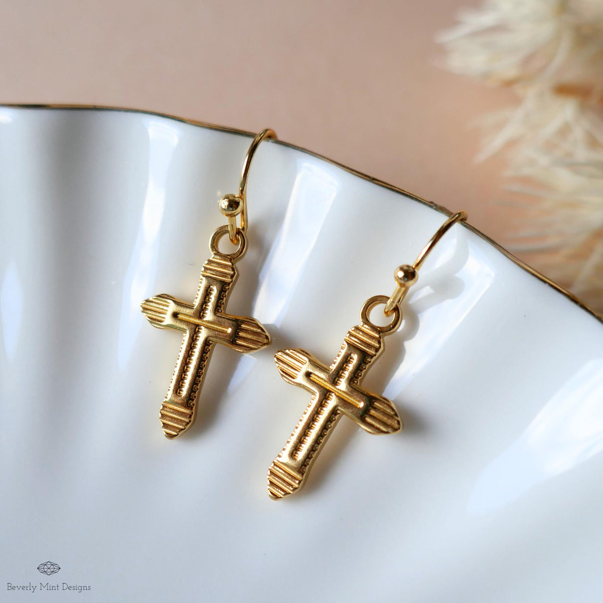 A pair of gold-colored earrings with a cross design, displayed on a white plate against a light pink background.