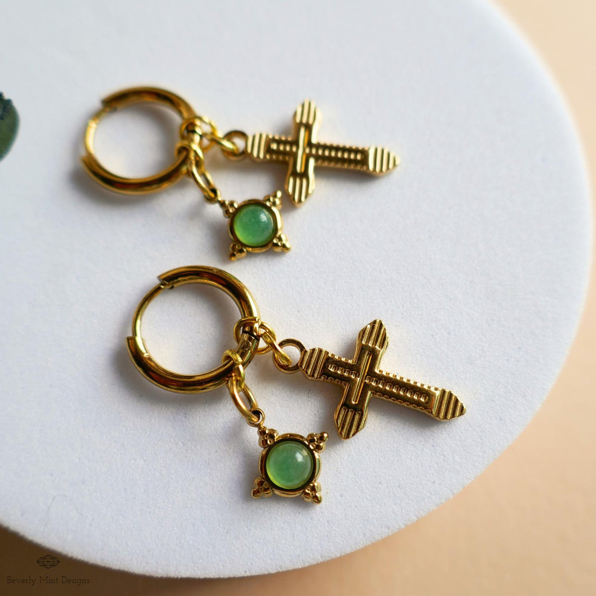 A pair of gold earrings with a cross-shaped charm and a green gemstone dangling from the earring hooks.