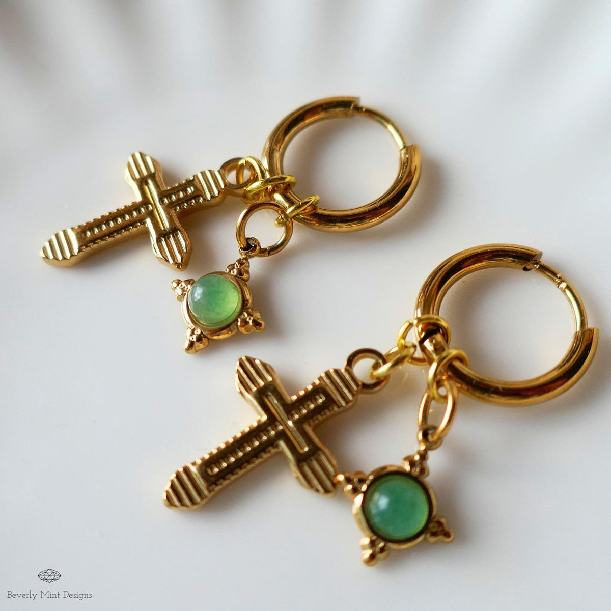 a pair of gold-colored earrings with green gemstones, including a cross-shaped charm and a round gemstone.