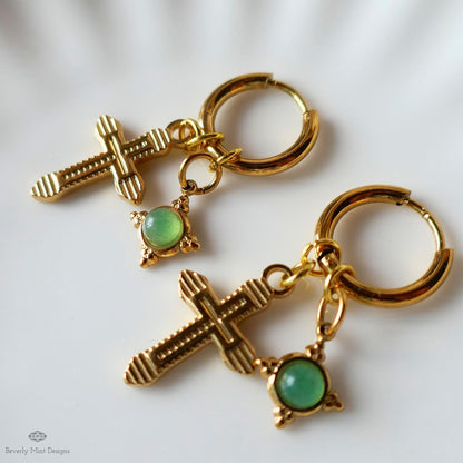 a pair of gold-colored earrings with green gemstones, including a cross-shaped charm and a round gemstone.
