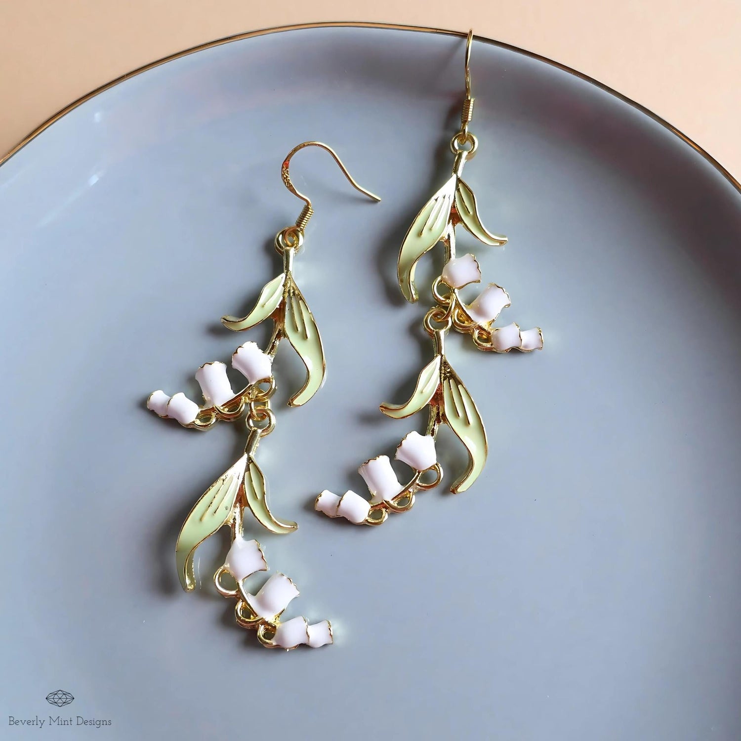 A pair of elegant earrings with white flowers and green leaves, displayed on a blue plate.