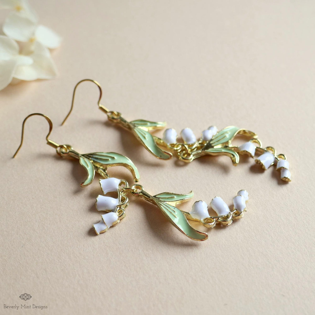 A pair of gold earrings with white flower-shaped charms and leaves, placed on a light-colored surface.