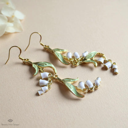 A pair of gold earrings with white flower-shaped charms and leaves, placed on a light-colored surface.