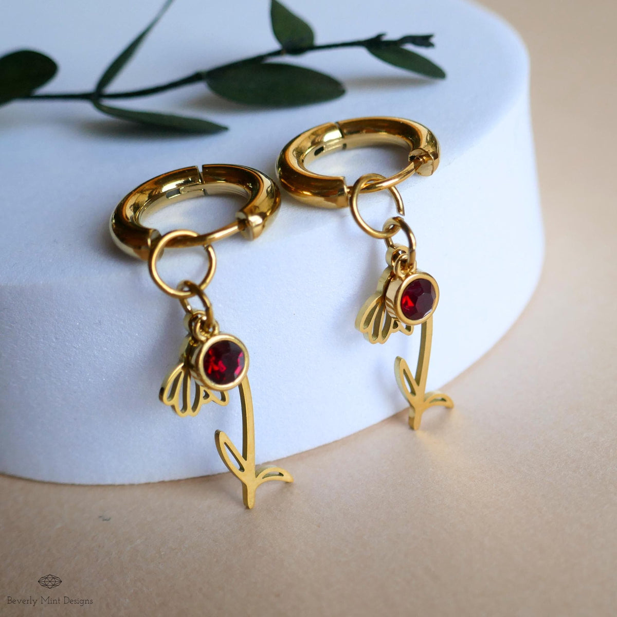 A pair of gold earrings with red gemstones and leaf-shaped charms, placed on a white surface.