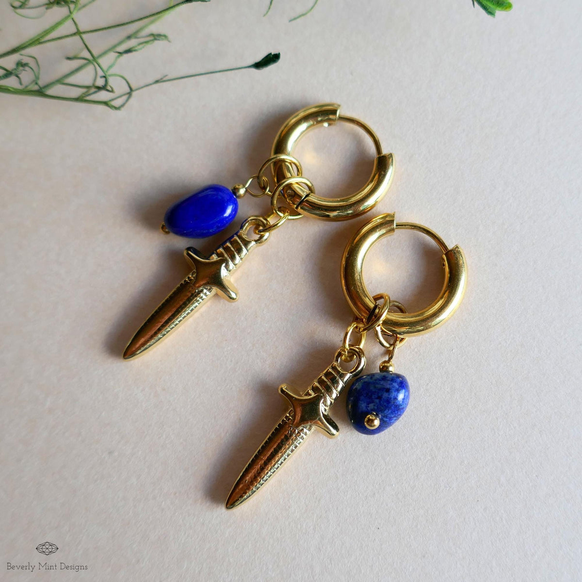 A pair of gold hoop earrings with a blue gemstone and a small blue gemstone dangling from them, placed on a white surface.