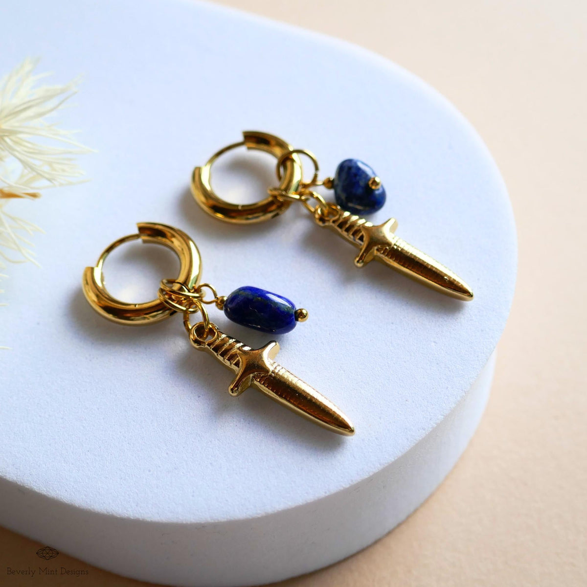 A pair of gold earrings with a knife-shaped pendant and blue gemstones, resting on a white surface.