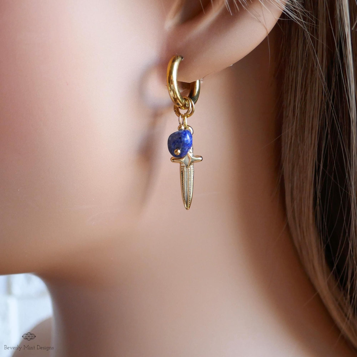 A close-up view of a person's ear wearing a gold earring with a blue gemstone and a small knife-like charm hanging from it.