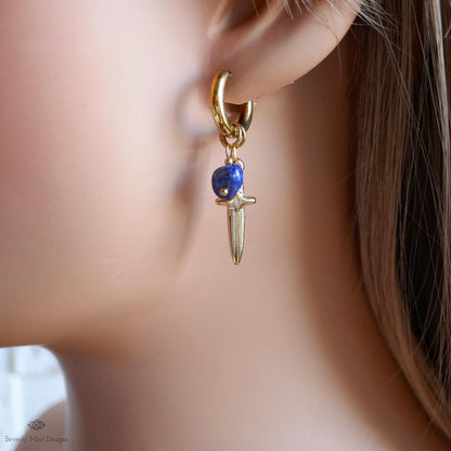 A close-up view of a person's ear wearing a gold earring with a blue gemstone and a small knife-like charm hanging from it.