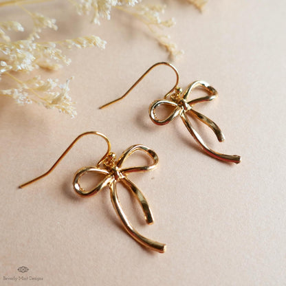 A pair of gold-colored earrings with a bow-shaped design, placed on a light-colored surface.