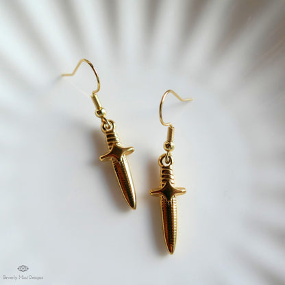 A pair of gold earrings with a sword-like design, placed on a white surface.