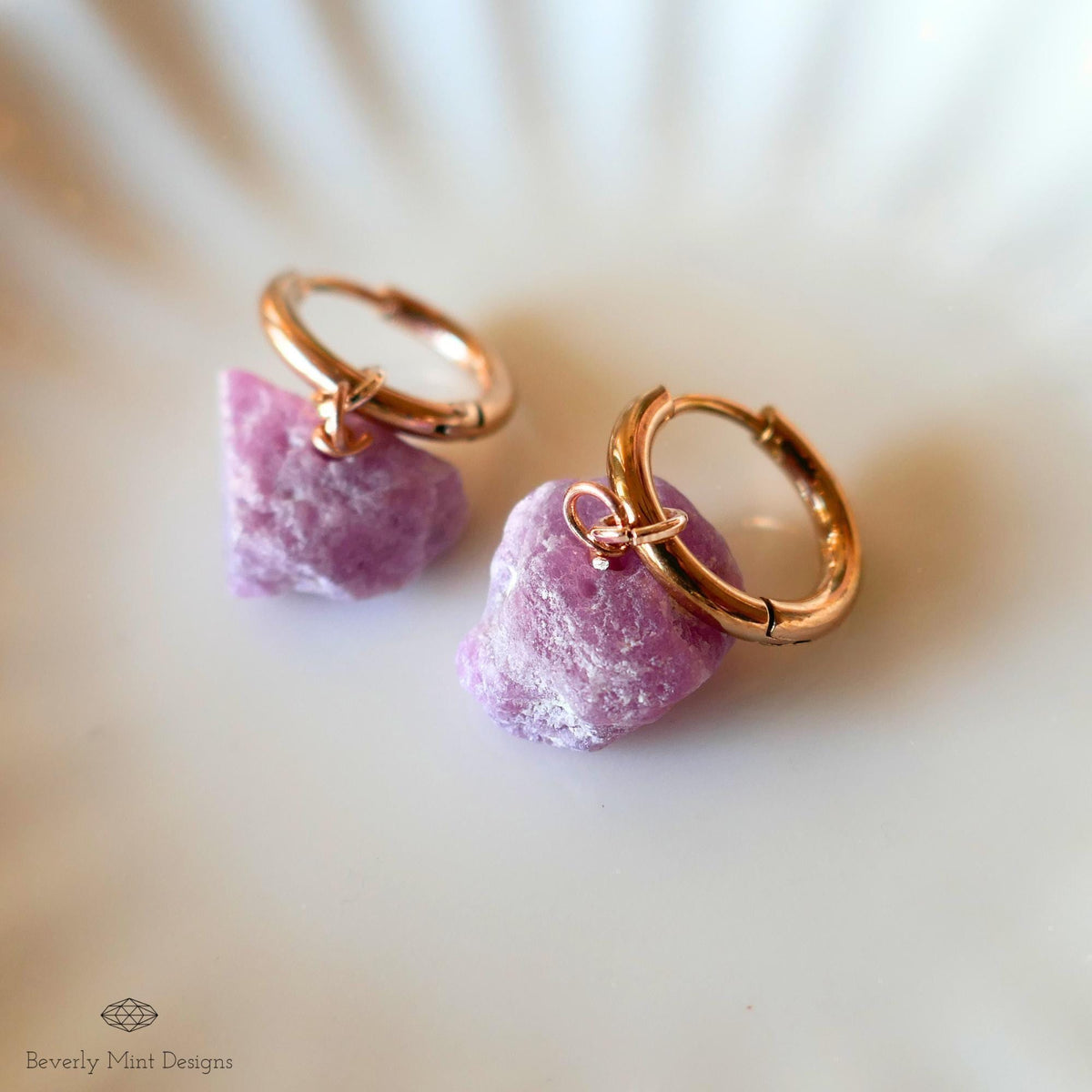 a pair of gold hoop earrings with a purple gemstone attached to them, resting on a white surface.