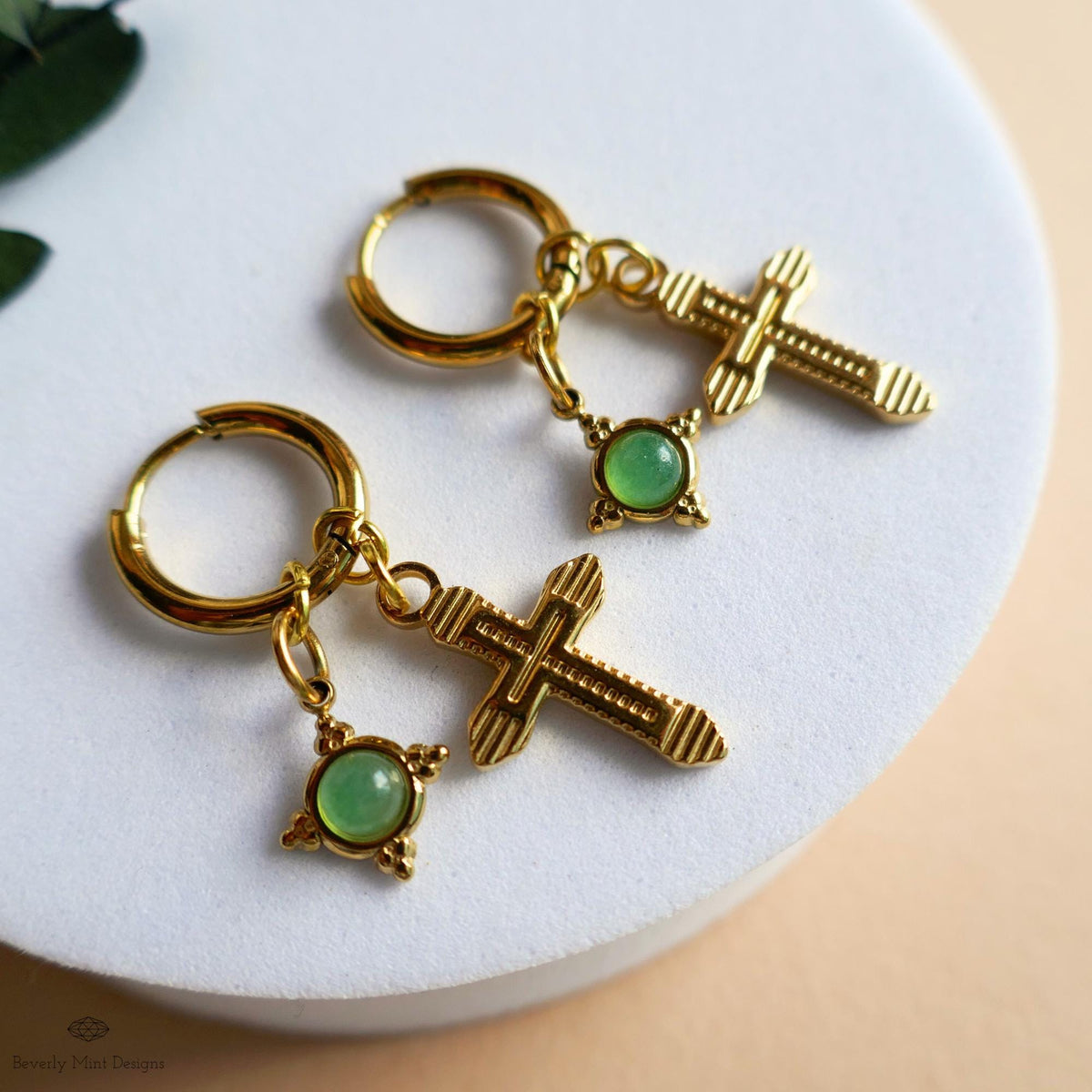 A pair of gold earrings with a cross-shaped charm and a green gemstone, resting on a white surface.