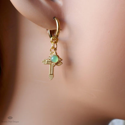 A close-up view of a person's ear wearing a gold-colored earring with a cross-shaped charm and a green gemstone.