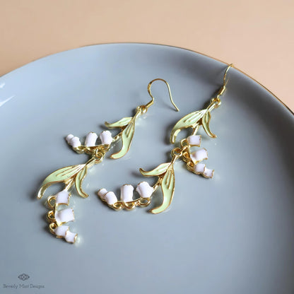 A pair of elegant earrings with white flowers and gold leaf-like designs, placed on a light blue plate.