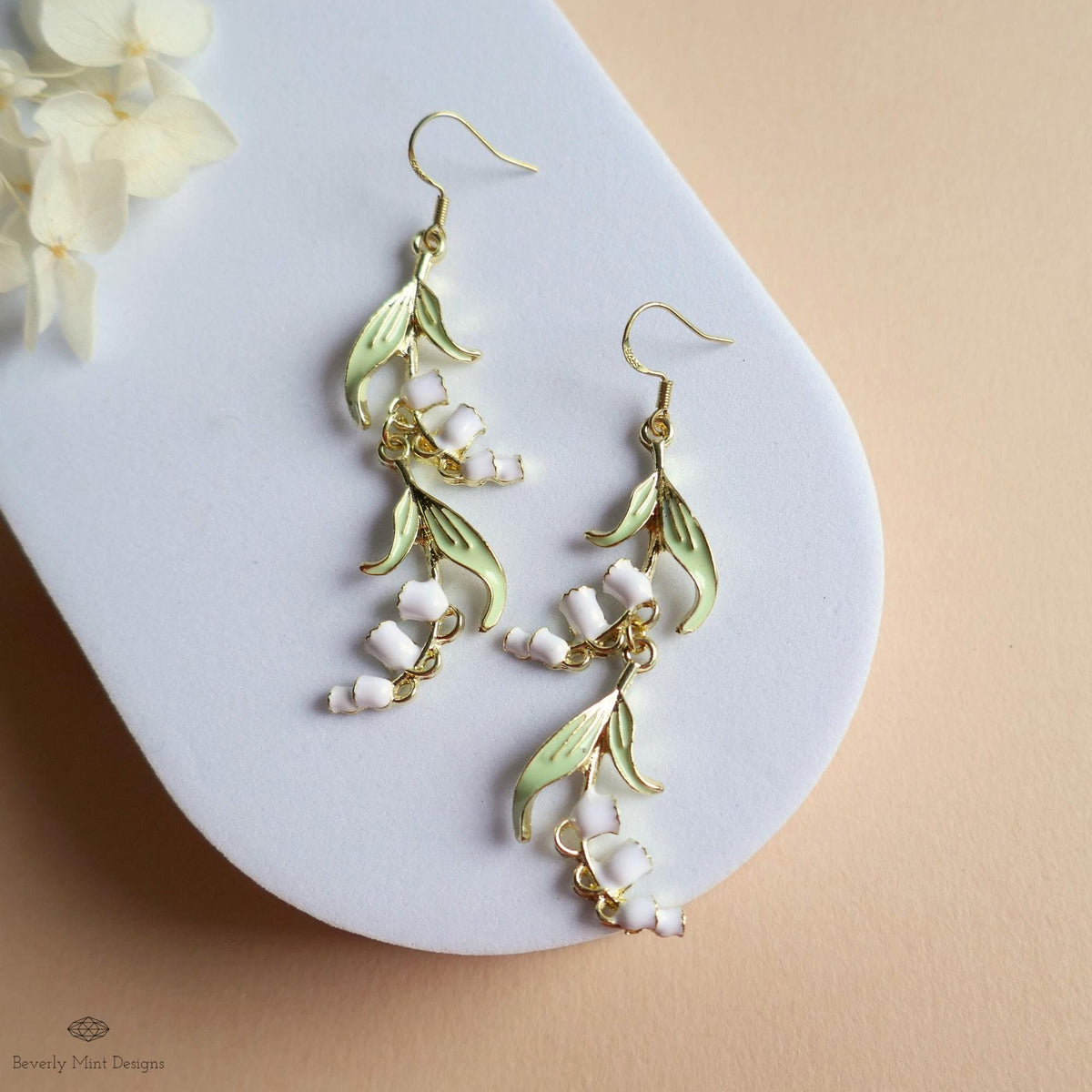A pair of elegant earrings with white flowers and green leaves, displayed on a white surface against a peach-colored background.