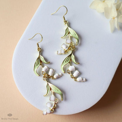A pair of elegant earrings with white flowers and green leaves, displayed on a white surface.