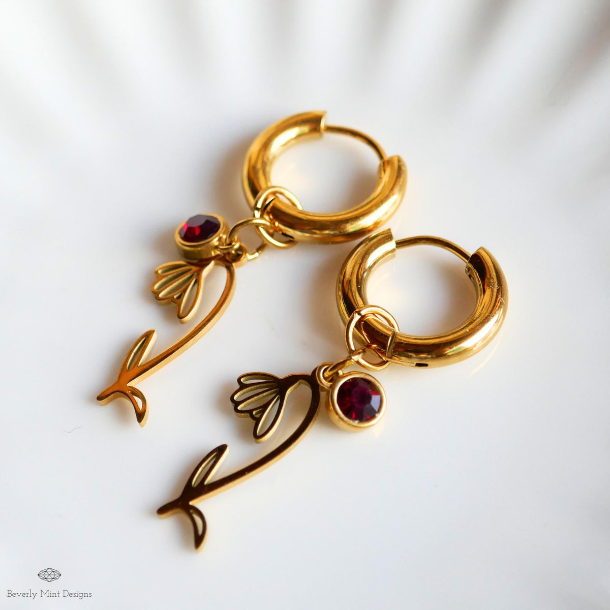 A pair of gold hoop earrings with red gemstones and floral charms, resting on a white surface.