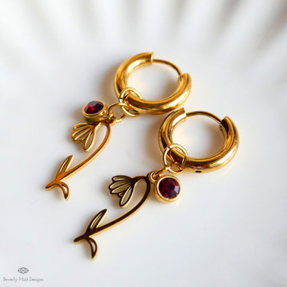 A pair of gold hoop earrings with red gemstones and floral charms, resting on a white surface.