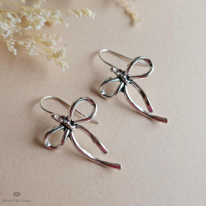 A pair of silver earrings with bow-shaped designs, placed on a beige background.
