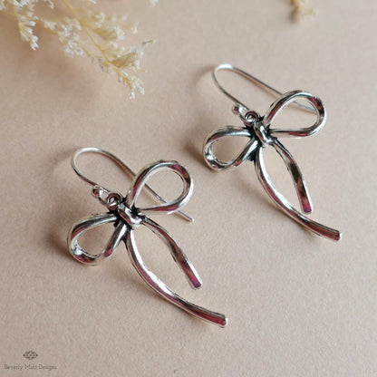 A pair of silver earrings with a bow-shaped design, placed on a beige background.