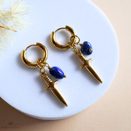 A pair of gold earrings with a blue gemstone and a small knife-shaped charm hanging from the earrings.