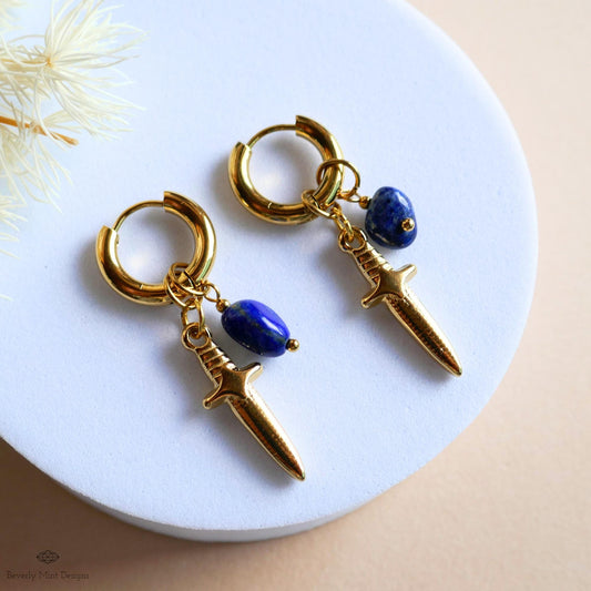 A pair of gold earrings with a blue gemstone and a small knife-shaped charm hanging from the earrings.