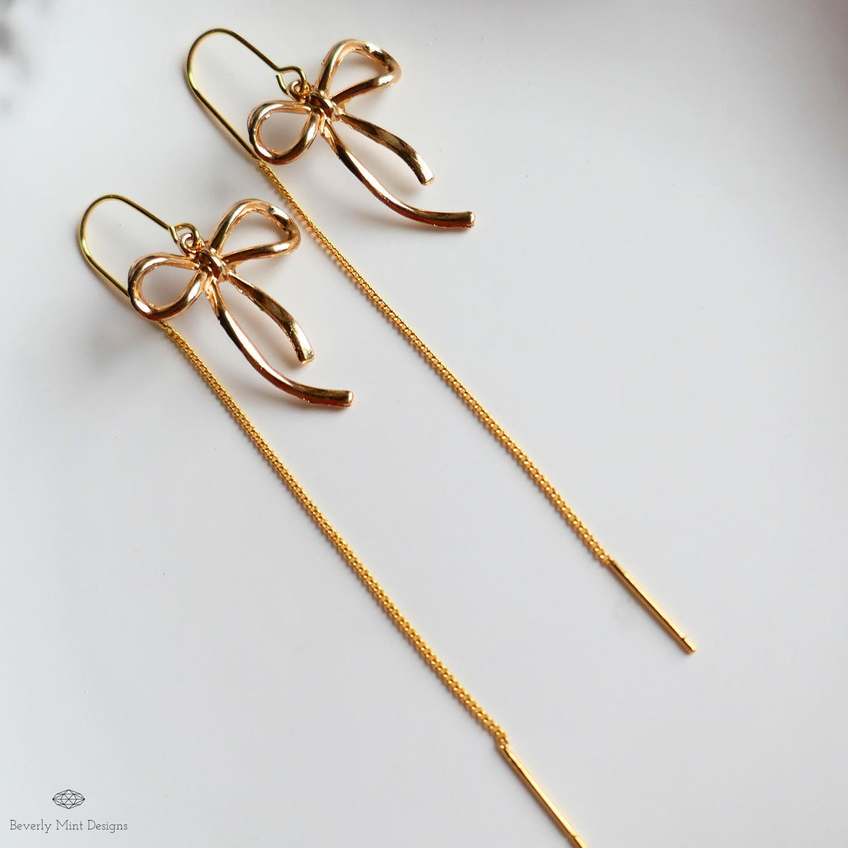 A pair of gold-colored earrings with bow-shaped designs, hanging from a chain.