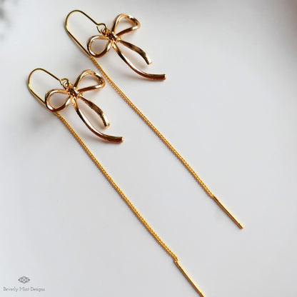 A pair of gold-colored earrings with bow-shaped designs, hanging from a chain.