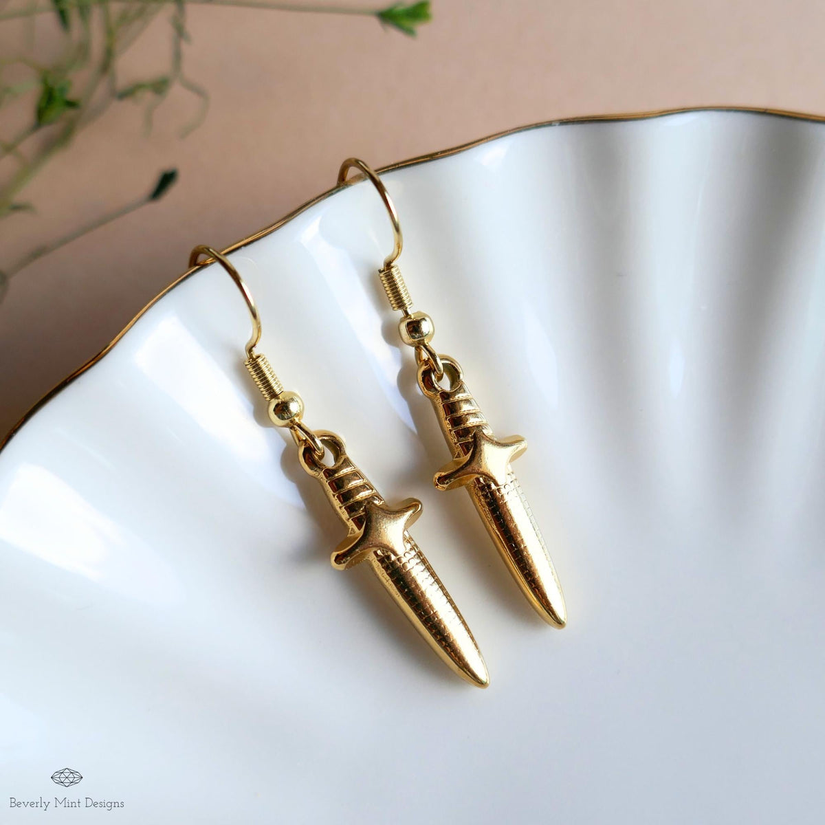 A pair of gold earrings with a knife-like design, placed on a white plate against a blurred background.