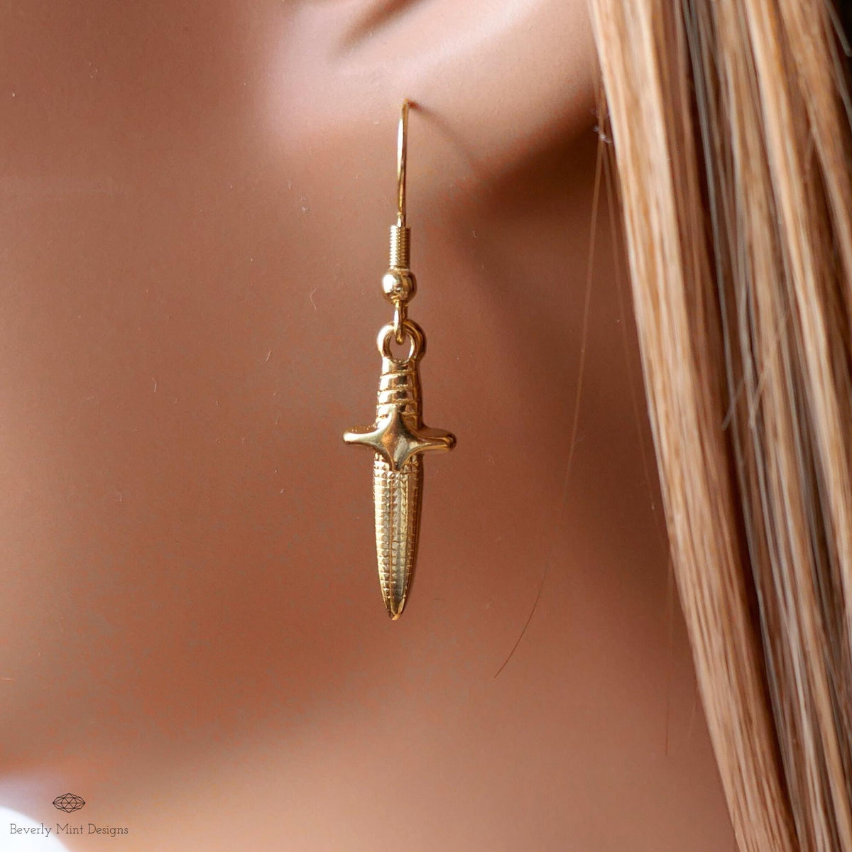 A gold earring with a cross-shaped pendant is shown against a blurred background.