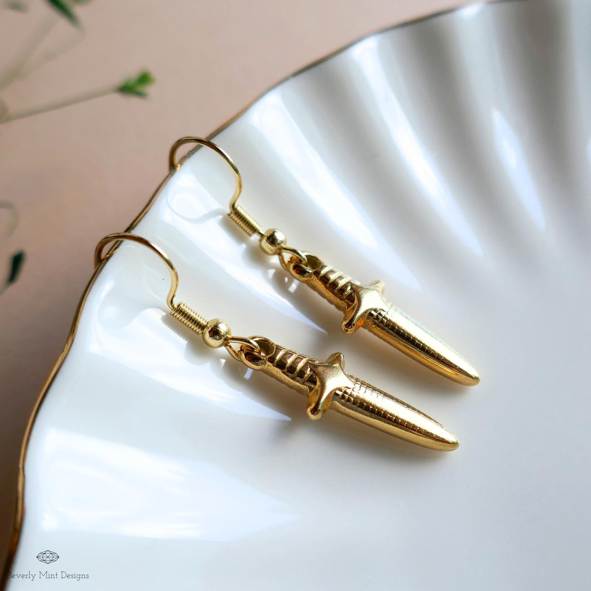 A pair of gold earrings with a knife-like design, resting on a white plate with a scalloped edge.