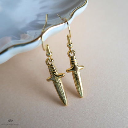 A pair of gold-colored earrings with a sword-like design, resting on a white surface.