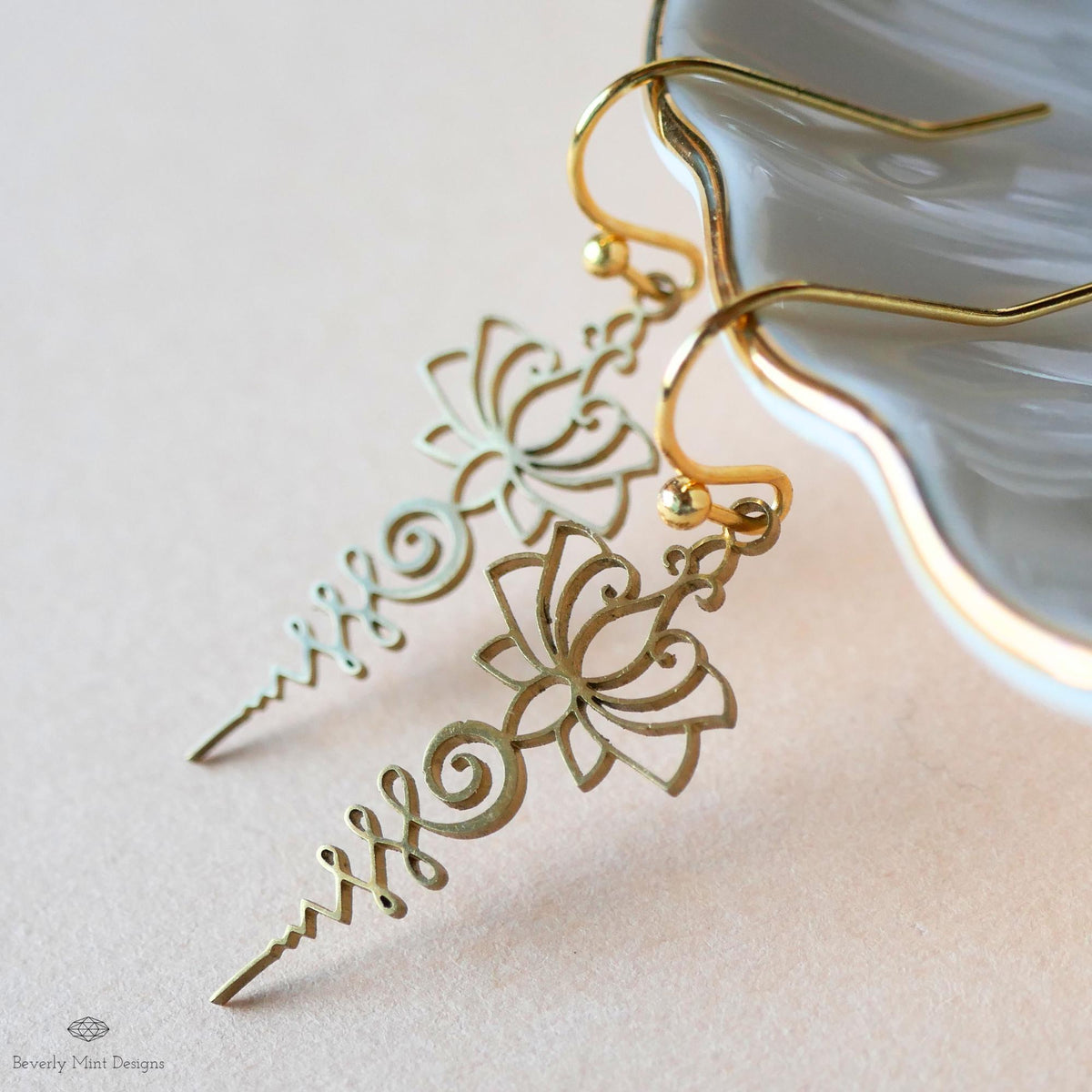 A pair of gold earrings with intricate floral designs, resting on a white surface next to a small white dish.