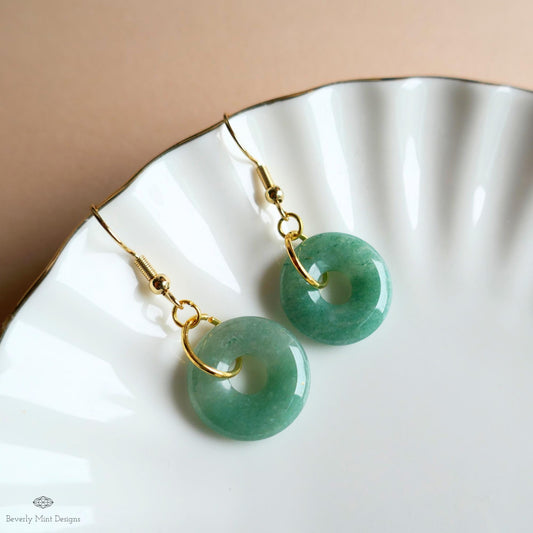 May include: A pair of jade donut earrings with gold-colored hooks. The earrings are a light green color and are displayed on a white, scalloped plate. The earrings have a circular design with a hole in the center.